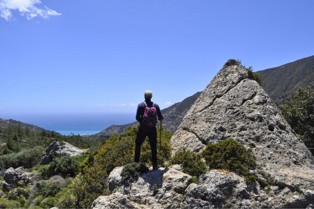 A hiker with a backpack stands on a rocky outcrop looking down at a traditional coastal village and the blue sea in a Self-Guided Hiking in Greece, Southwestern Crete.